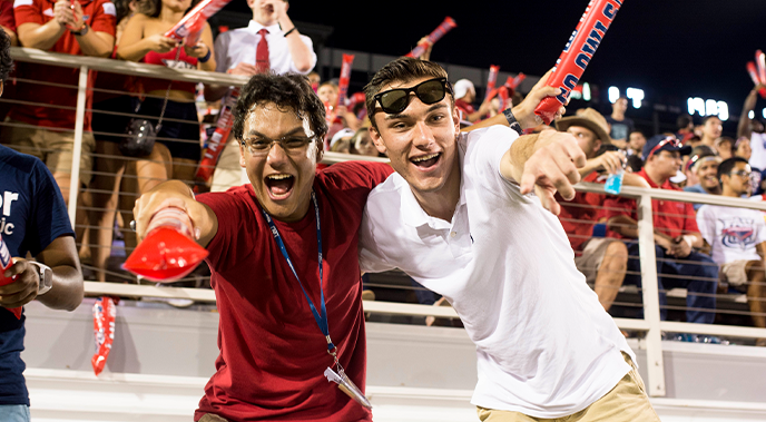 Two male students during an 鶹Ů football game smiling and pointing at the camera